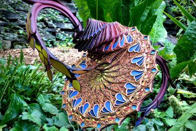 Similarity of Being is an artwork made from cut paper, wood, gold leaf, botanicals, water, and PVC. It looks like an exotic flower - blue and purple petal shapes fan around a circular base, while a piston-looking structure rises from the top.