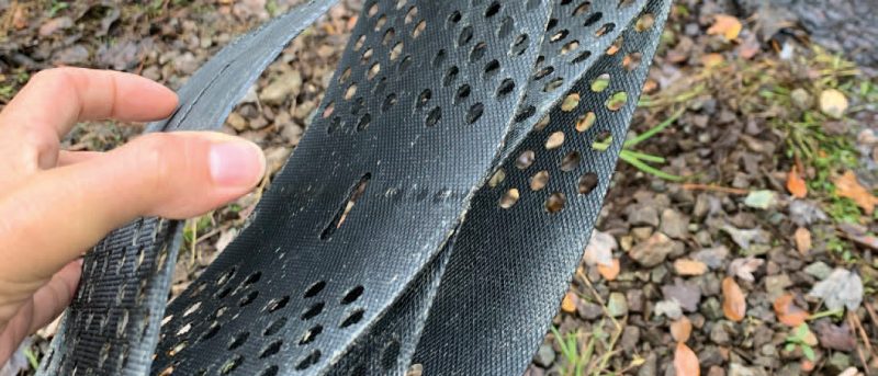 Close up of a hand holding a Geoweb textile. The textile is a gray compressed hexagon made of hard plastic with small round holes in the sides. In the background there is a small woods stream.