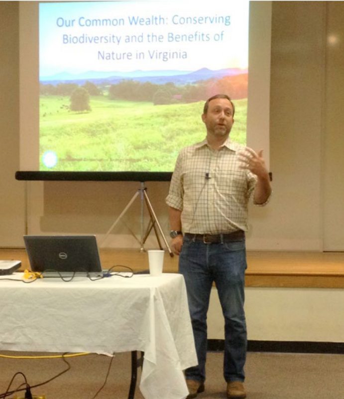 A speaker stands behind a table and in front of a slide presentation.