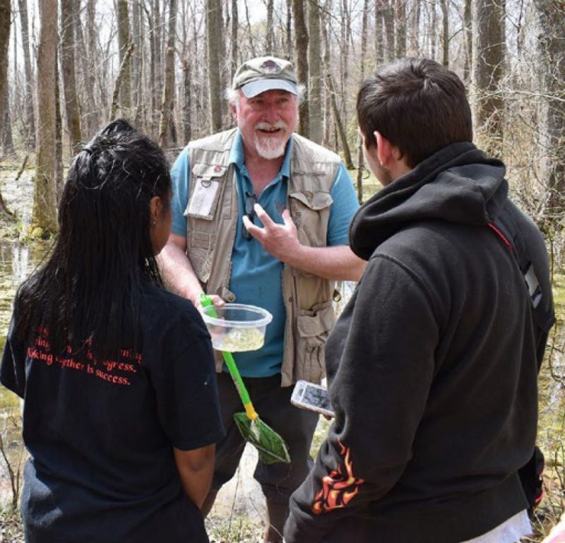 A man holding a water skimmer and a plastic container, talks with two young people who have their backs to the camera 