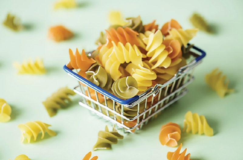 A small basket of creamy white, pale green and light orange rotini pasta in a small wire basket with other rotini noodles scattered on the surface all around the basket.