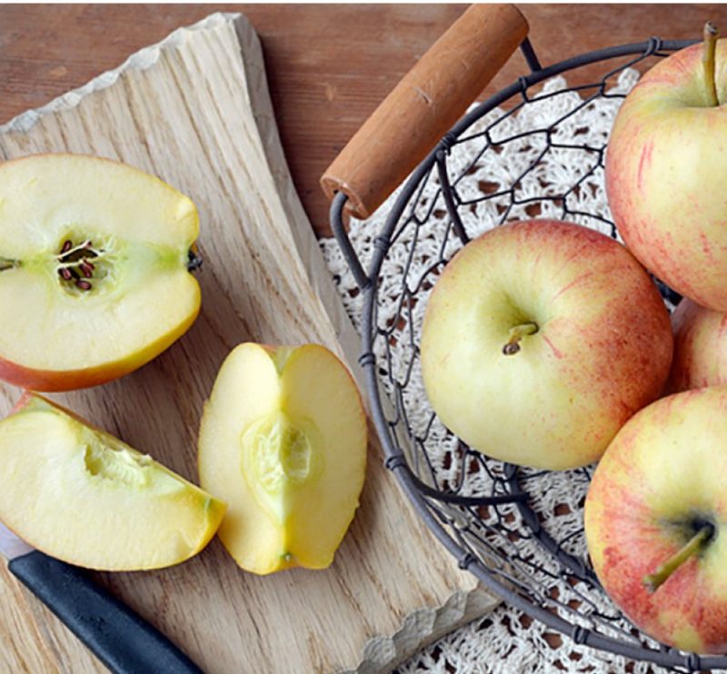Overhead shot of a decorative colander holding four apples next to a cutting board with one apple that has been cut into three pieces.