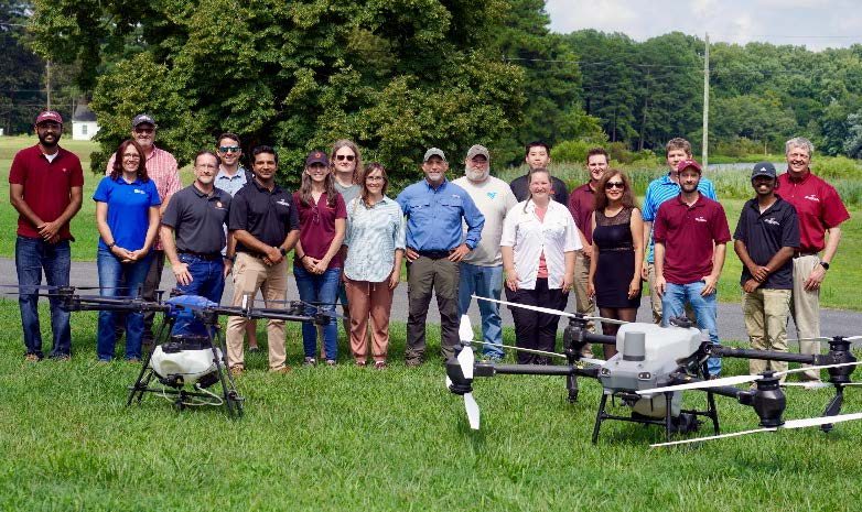 Participants of Mid-Atlantic spray drone workshop standing behind two large drones in a field.